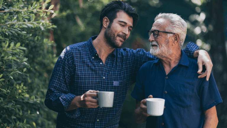 A younger man and an older man stand outdoors in a leafy garden, smiling at each other with their arms around one another’s shoulders, each holding a white mug.